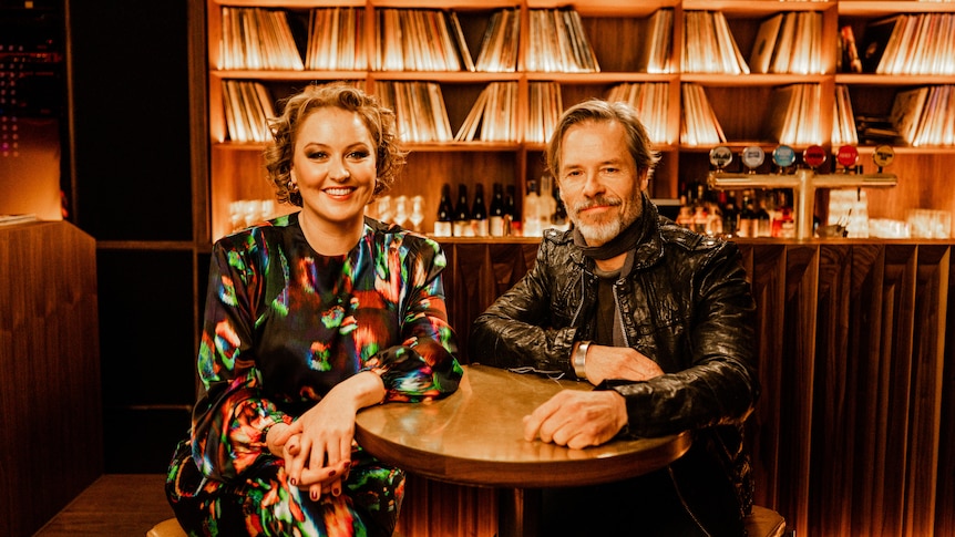 Zan Rowe and Guy Pearce sit at a table in a bar with ambient lighting and vinyl records behind them