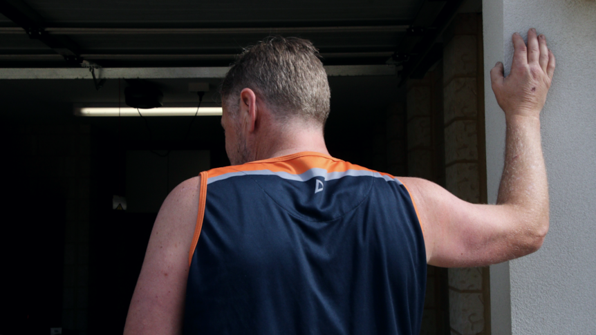 A man with short fair hair, wearing a sleeveless shirt, stands with his back turned and his right arm raised up to a wall.