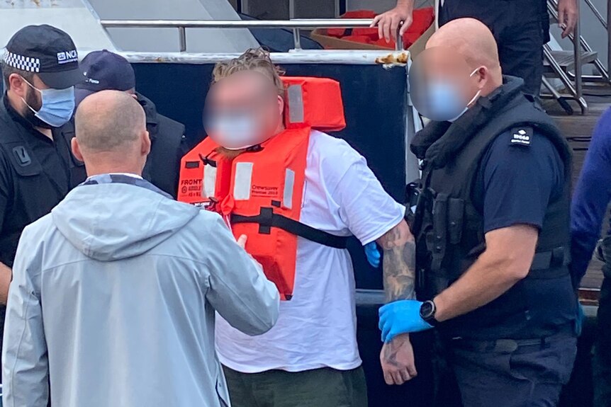 A man wearing a life jacket and with his face blurred is led away by several police officers.