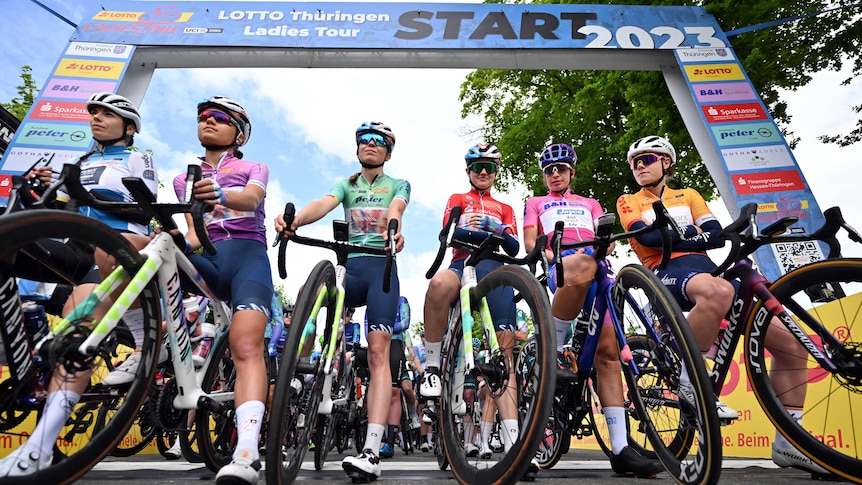 Women in lycra and helmets in racing gear line up at starting line for road cycling race.