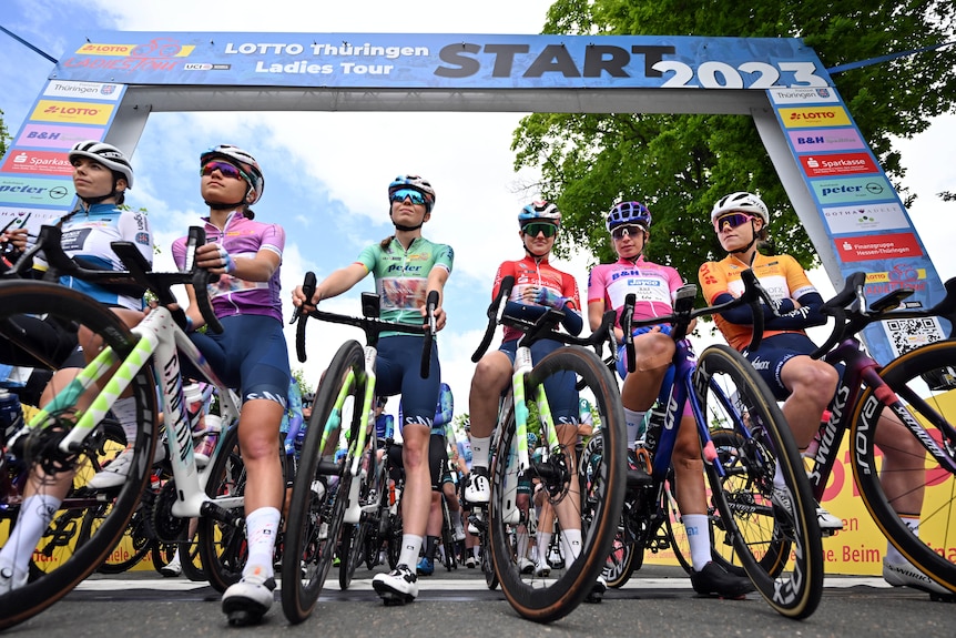 Women in lycra and helmets in racing gear line up at starting line for road cycling race.
