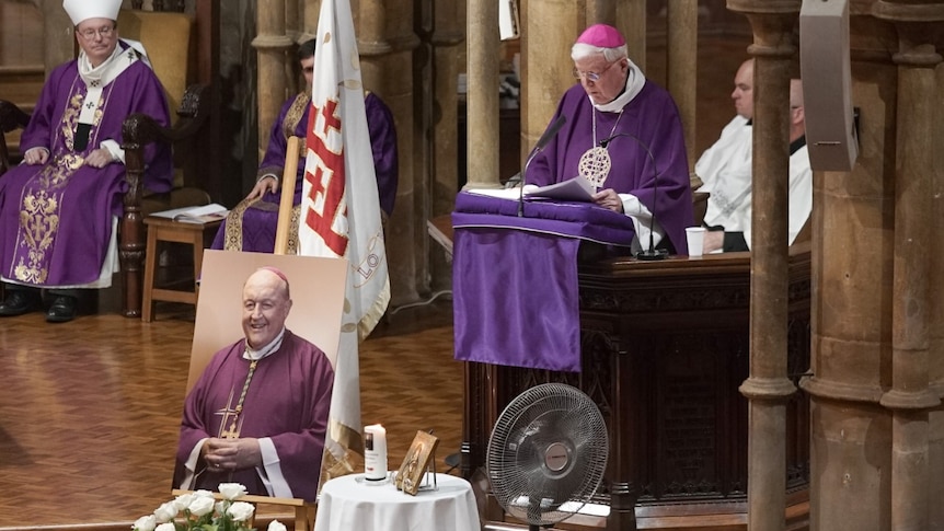 A man speaks from behind a portrait of former Archbishop Philip Wilson