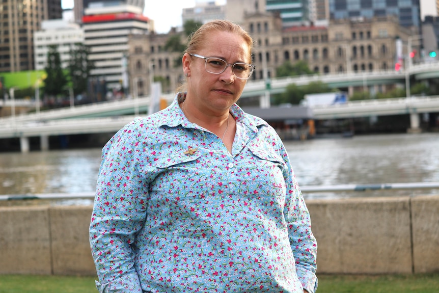 A woman wearing a floral shirt and a bee brooch stands in front of a river, looking serious.