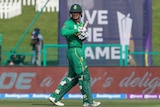 A cricketer wearing an all green uniform and helmet walks from the field carrying a bat. 