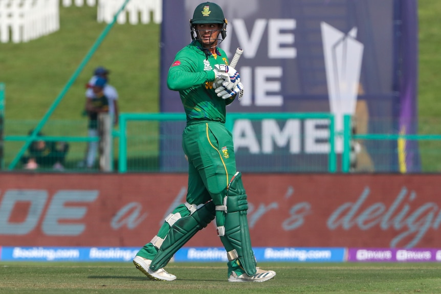 A cricketer wearing an all green uniform and helmet walks from the field carrying a bat. 