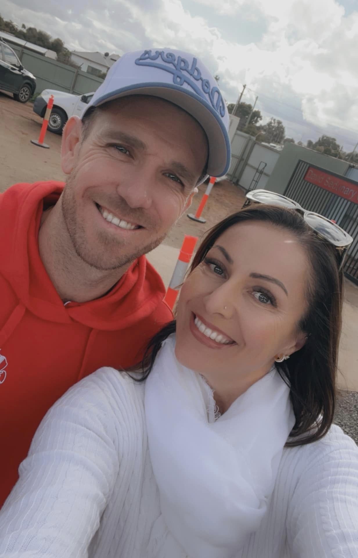 A man wearing a red jumper and a woman wearing sunglasses smiling in a selfie in front of a canteen