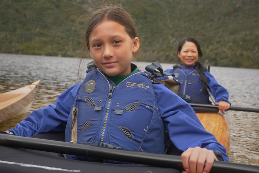 A girl with paddle in hand, sitting in the front of a kayak, her mother behind, on a lake with mountains behind.