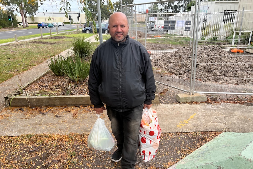 Imad standing on a footpath, holding a bag of groceries in each hand.