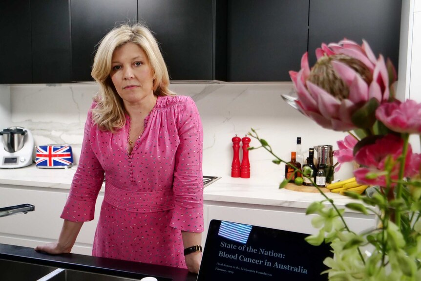 Deborah Sims, a blonde woman in a pink top, looks at the camera in her kitchen.