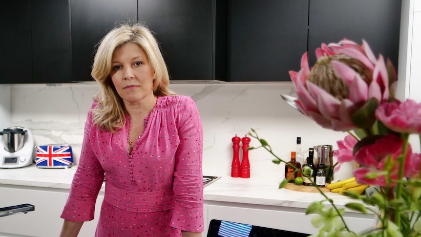 Deborah Sims, a blonde woman in a pink top, looks at the camera in her kitchen.