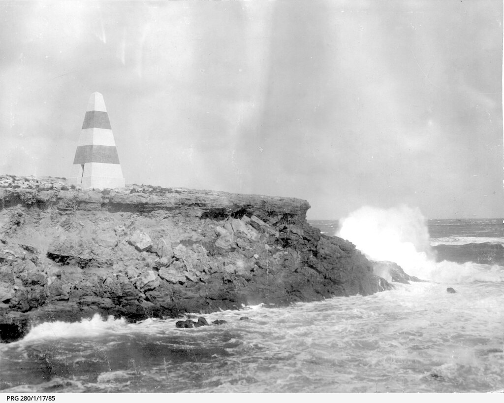 A black and white photograph of a cliff with a striped pyramid-shaped obelisk atop it, and waves crashing into it