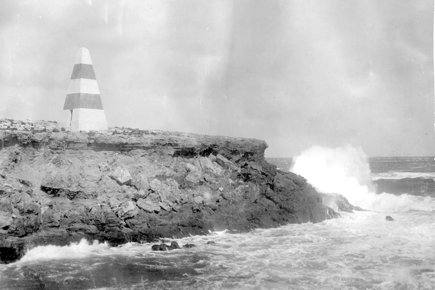 A black and white photograph of a cliff with a striped pyramid-shaped obelisk atop it, and waves crashing into it