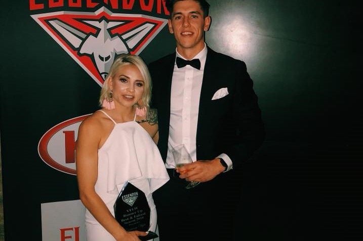 Ugle and her boyfriend pose in front of an Essendon logo, dressed in black tie, Ugle holding an award.