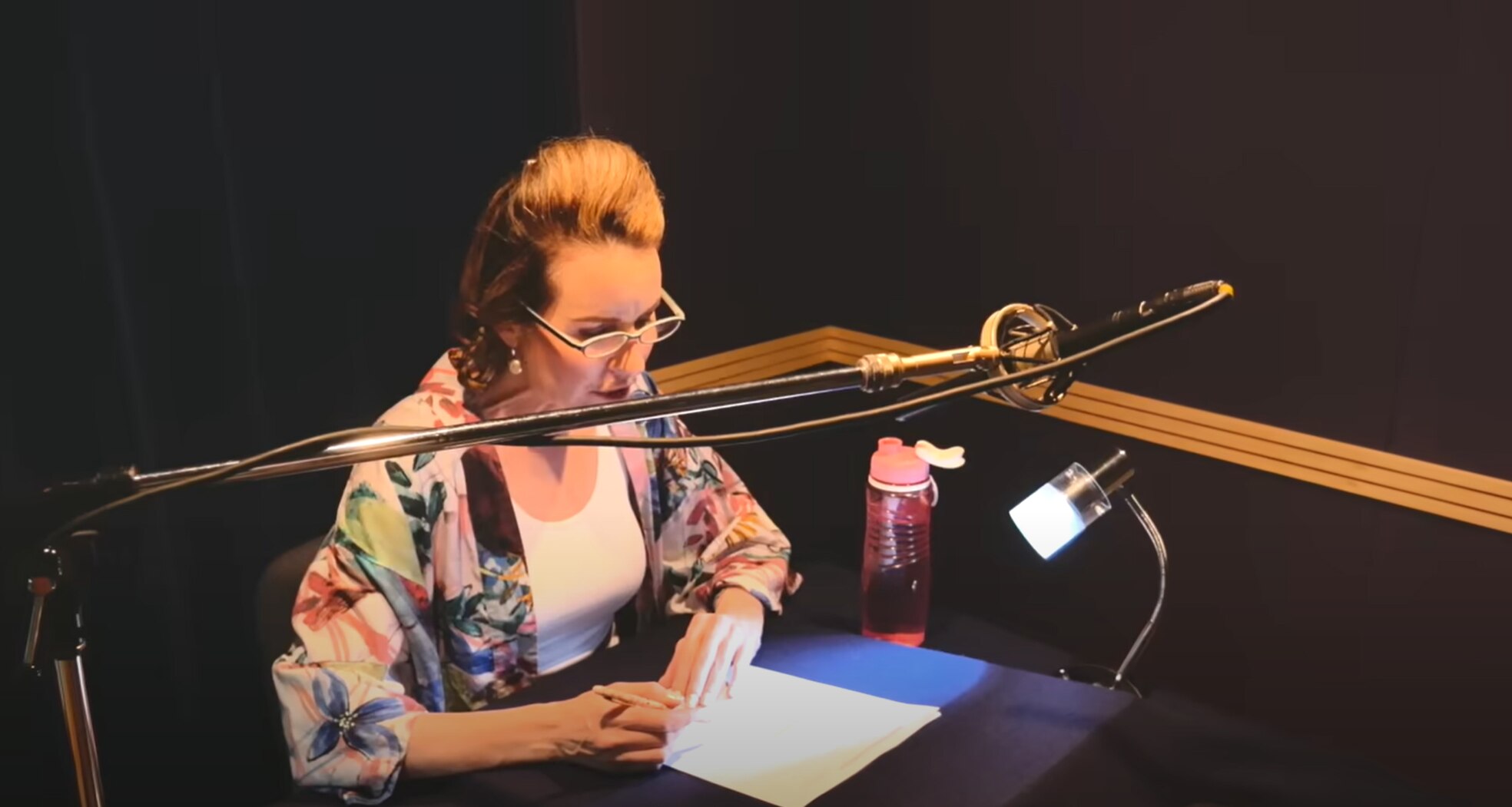 a woman sits at a desk and reads off a sheet of paper while speaking into a microphone
