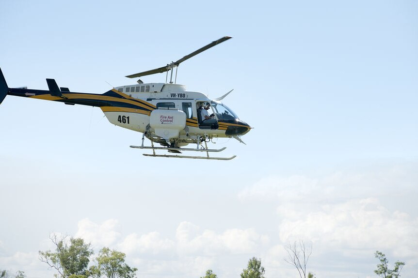 A helicopter with a container on the side reading 'Fire Ant Control' flies low over treetops.