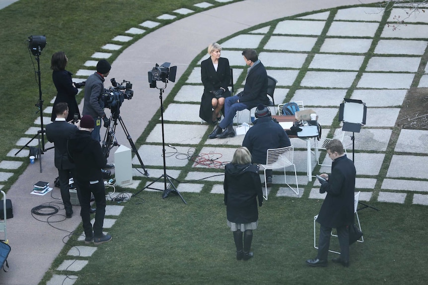 Wide shot Rowland and Bishop sitting on chairs outside surrounded by camera, lights and production crews.