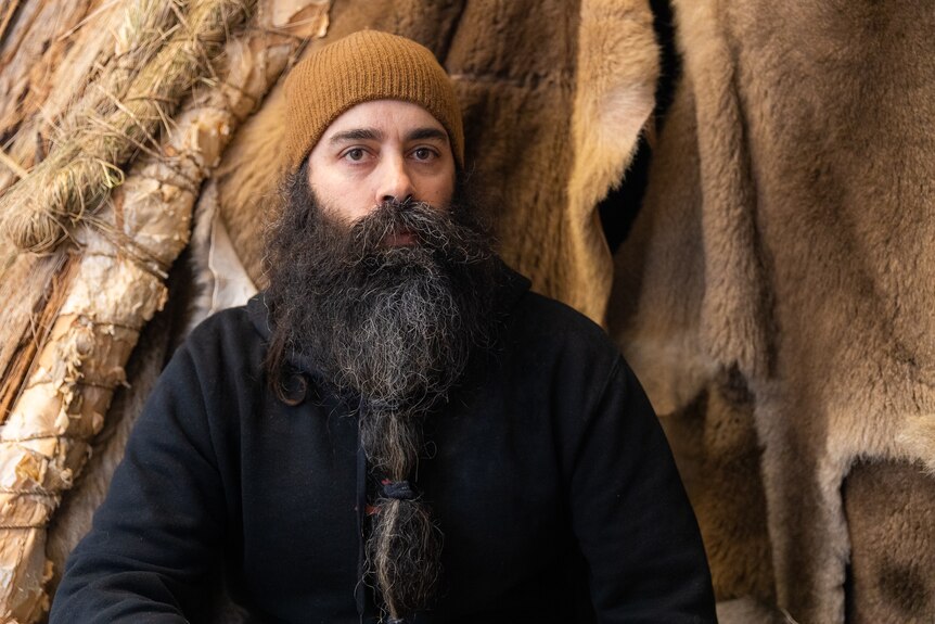 An Indigenous young man with a big beard and a beanie sitting in front of animal skins and bark in an artwork at Dark Mofo