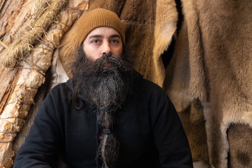 An Indigenous young man with a big beard and a beanie sitting in front of animal skins and bark in an artwork at Dark Mofo
