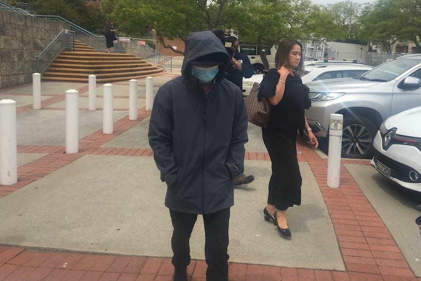 A man in a hooded navy jacket and mask walks past reporters alongside a woman in a black dress.
