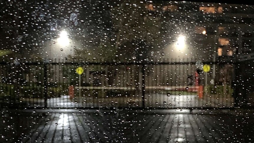 Streetscape at night, rain drops in the foreground, in front of an industrial fence, boom gate and then lights in distance.