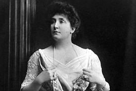 Black and white photo of Dame Nellie Melba standing holding a long pearl necklace.