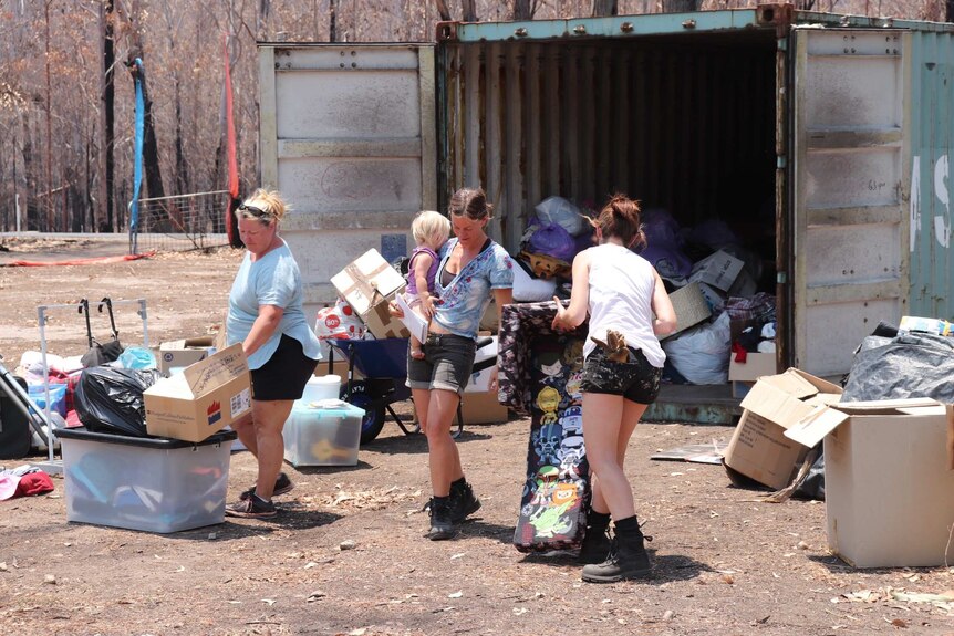 Three women carry goods, one also carries a toddler, from a shipping container loaded with bags and boxes, on a charred property