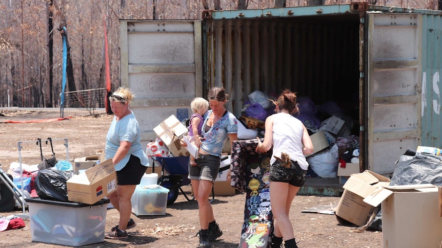 Three women carry goods, one also carries a toddler, from a shipping container loaded with bags and boxes, on a charred property