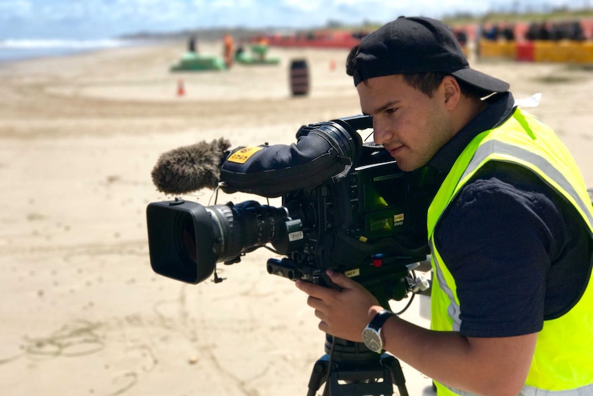 Haidarr wearing baseball cap and green high vis vest looking down camera viewfinder at beach.