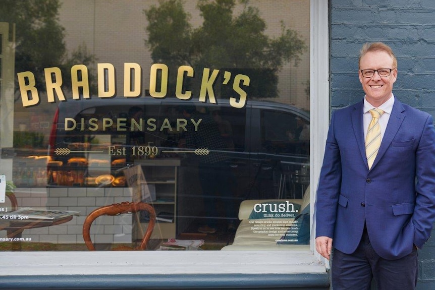 A wide shot of Bruce standing next to a shop front window with words saying 'Braddock's dispensary'.