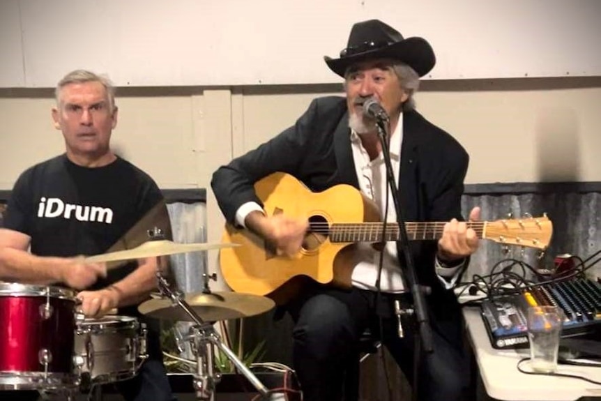 A man wearing a black hat and jacket strumming a guitar and singing while another man wearing a black shirt plays drums