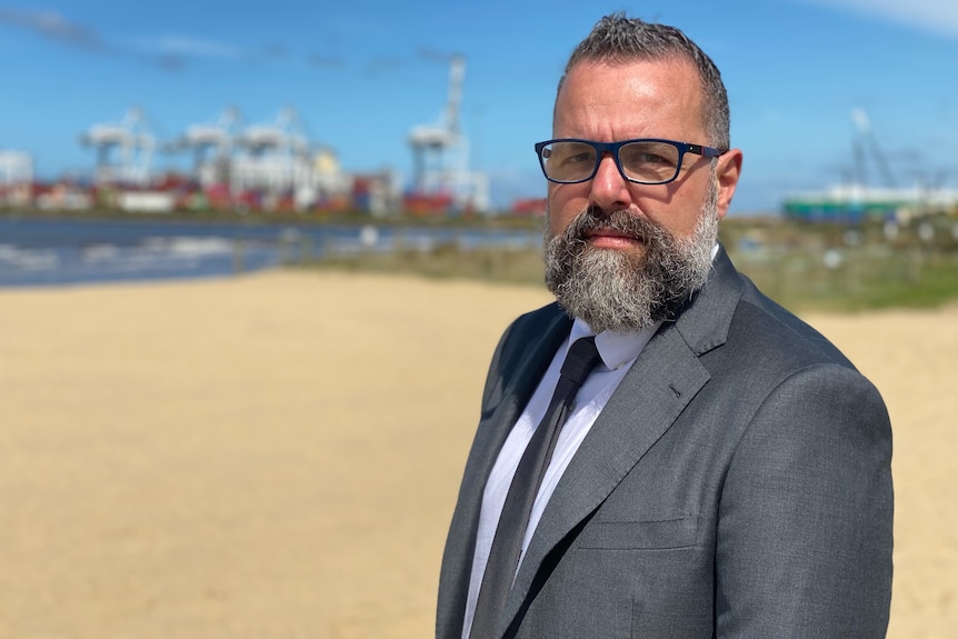 A man in a grey suit with grey hair and beard and glasses standing in front of a shipping port.