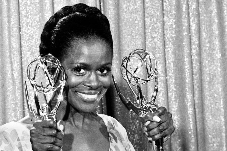 Cicely Tyson smiles and looks to the camera as she holds two golden Emmy Awards in a black and white photo against a curtain.