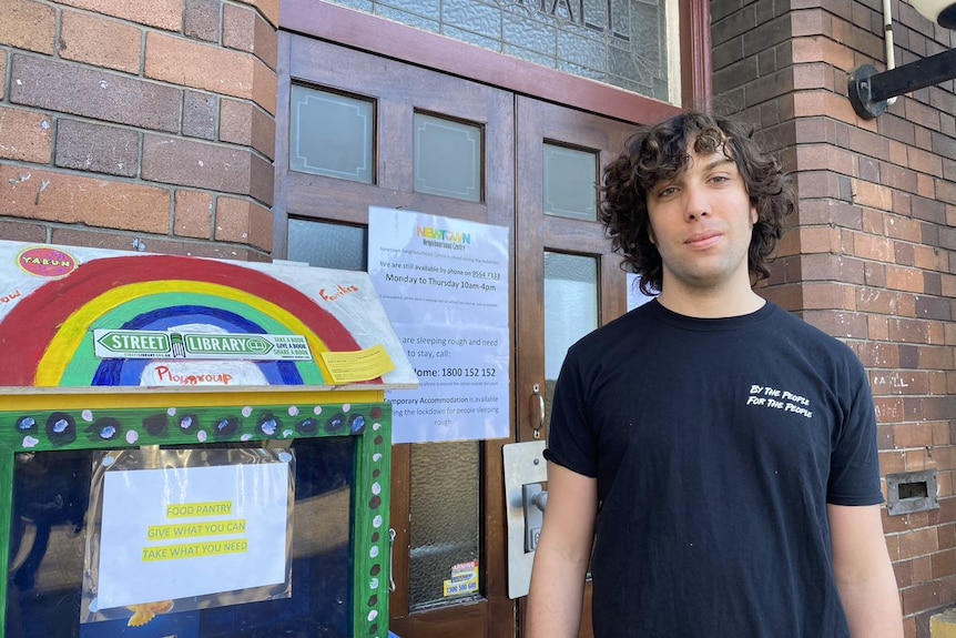 A young man with curly hair stands in front of a community centre.