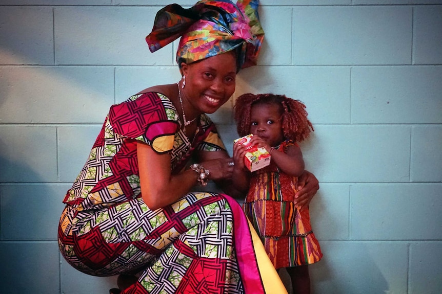 An older lady wearing a head-dress crouches down to pose with a little girl drinking from a juice box.