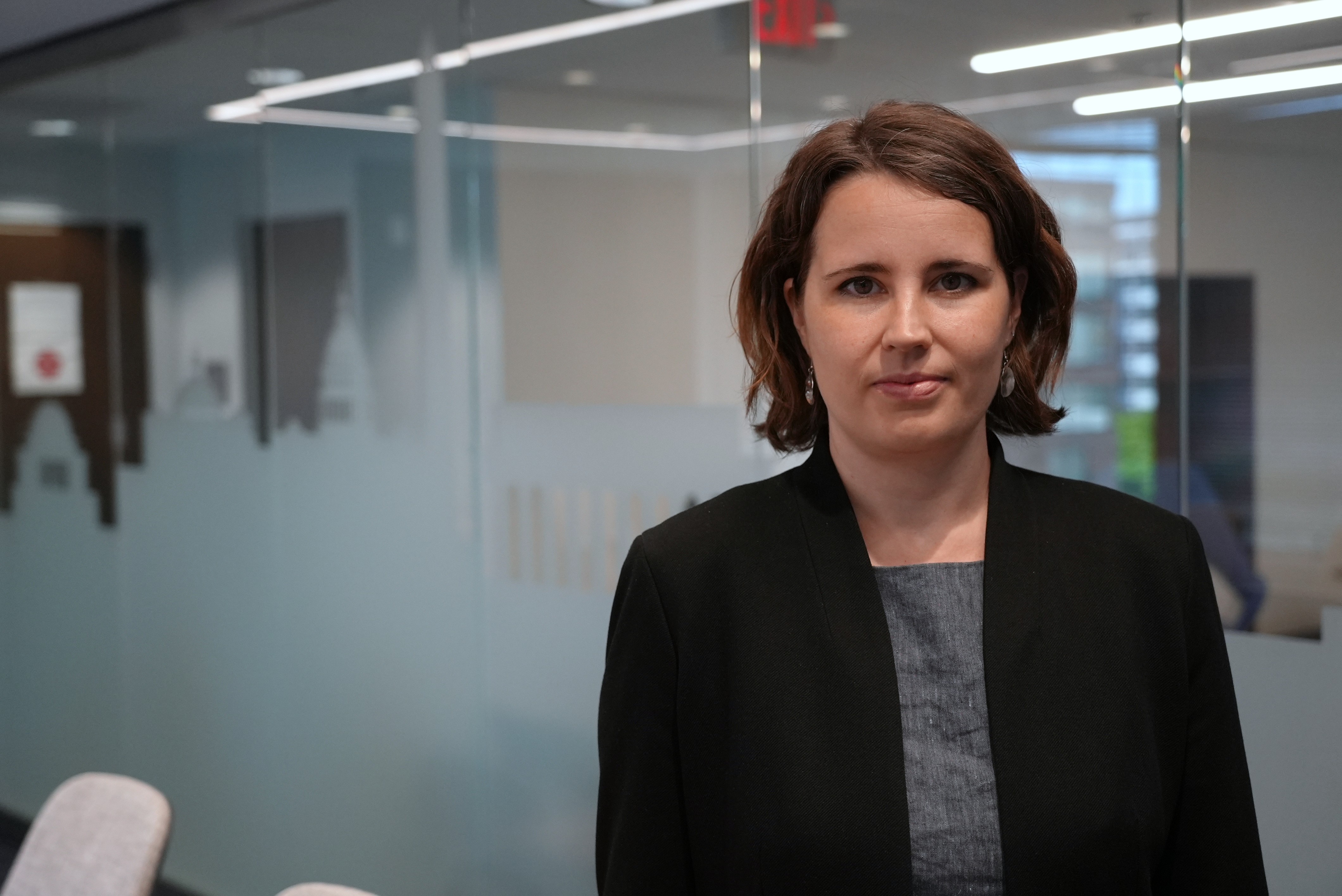 A woman with a brunette bob wearing a black blazer stands in a nondescript office with a neutral facial expression.