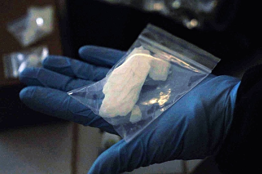 A blue-gloved hand holds a small plastic bag containing a large white crystal.