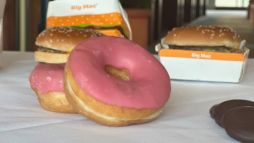 A donut with pink icing, leaned up against another donut with two big mac burgers behind it.