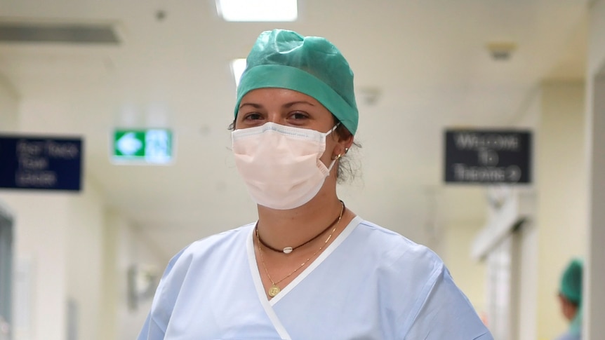 A woman smiling in scrubs and wearing a mask in the hallway 