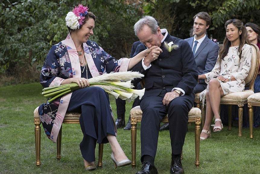 A couple (one in their 40s, one in their 60s) sit in fashionable formal garb, the woman holding a bouquet of lilies.