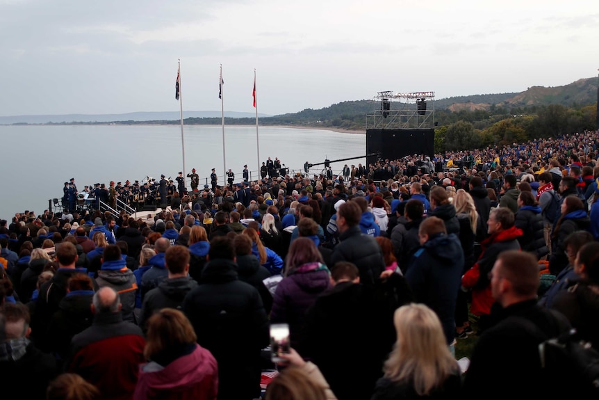 A large crowd of people gather at dawn by the water in front of three flag poles