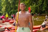 A young shirtless guy grinning and gripping an oar on a river bed 