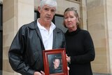 A man wearing a black leather jacket and a woman wearing a black jumper stand holding a framed photo of their son.