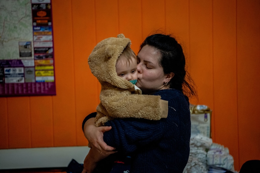 A woman holds and kisses a baby, red wall in background.