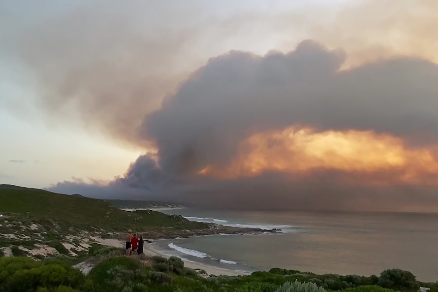 Three people stand on a remote beach looking at thick smoke from a bushfire filling the horizon in the distance.