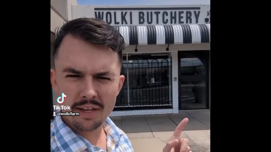 A man stands in front of a butcher shop pointing at the sign which reads Wolki Butchery