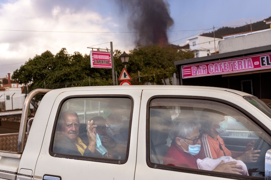 Four elderly people sit in a car as black smoke rises in the background