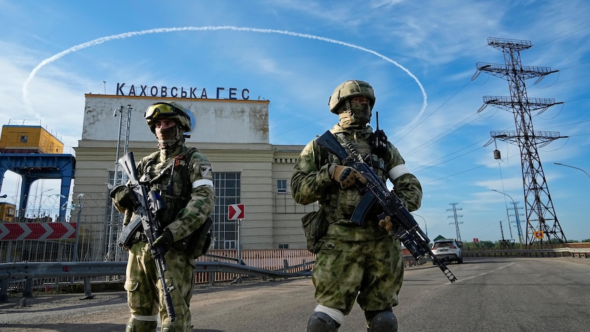 Two armed Russian soldiers guard an entrance of a rundown hydroelectric station, with powe lines running behind them.