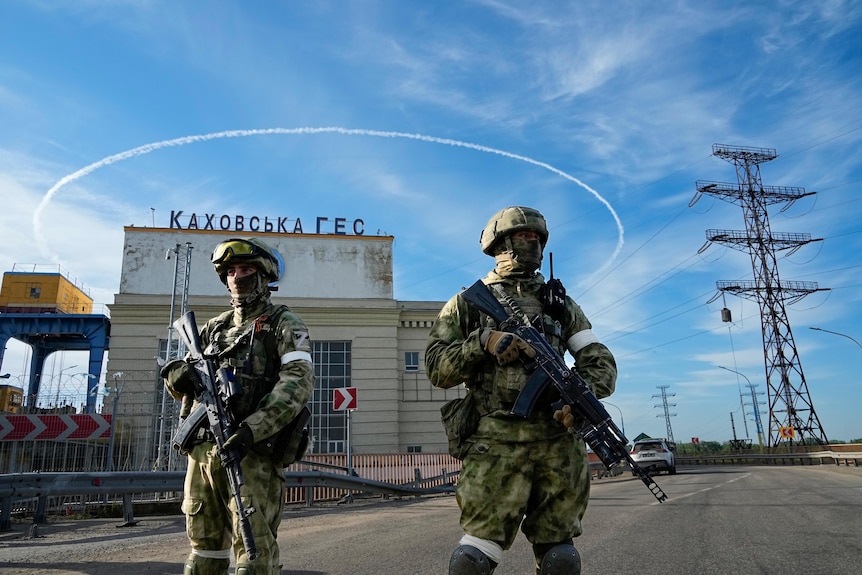 Two armed Russian soldiers guard an entrance of a rundown hydroelectric station, with powe lines running behind them.