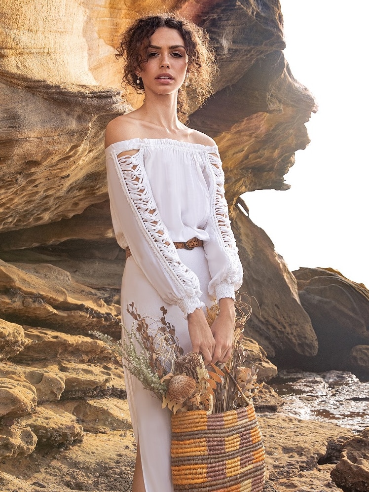 A woman in a white dress and a woven bag sands near rocks on a beach.