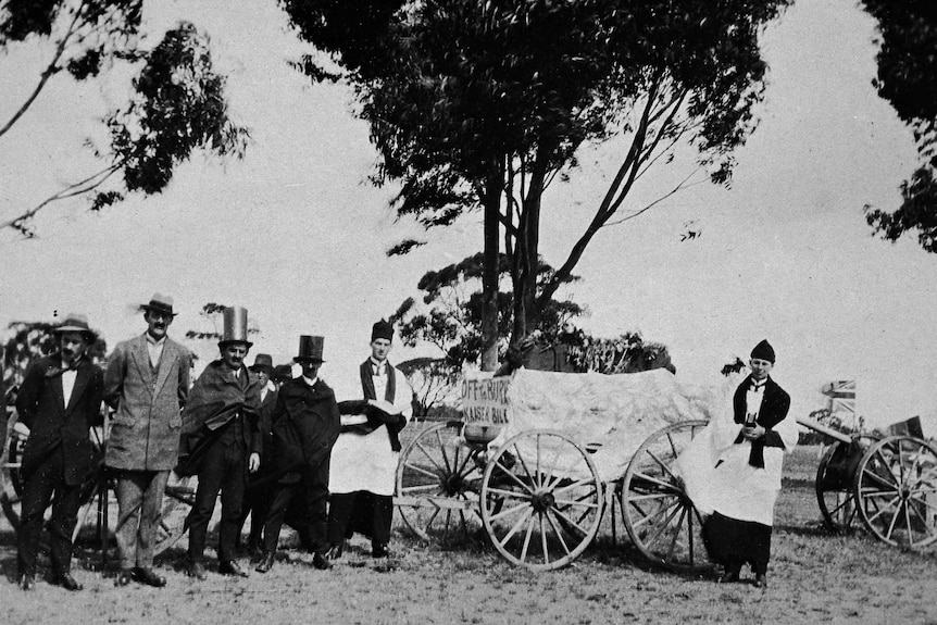 Seven men standing outdoors around a funeral card with a sign reading 'Off to bury Kaiser Bill'.